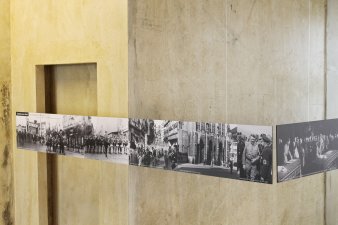 Zwei lange Tafel hängen im 90 Grad Winkel zueinander in einem Raum mit gelben Wänden. Auf den Tafeln sind Schwarz-Weiß-Fotografien zu sehen, die Stadtansichten mit Menschenansammlungen zeigen.