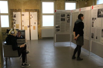 In der rechten Bildhälfte steht eine junge weibliche Person vor Stehtafeln mit Schrift und Bildern. In der linken Bildhälfte sitzt eine junge weibliche Person mit Kopfhörern vor einem Tisch, auf dem ein Monitor steht. Der Monitor zeigt Kopf eines älteren Mannes. Im Hintergrund sind drei menschengroße Tafeln an die Wand gelehnt.