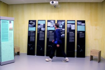 Links eine Vitrine, mittig stehen sechs Ausstellungstafeln, ein junger Mann läuft an ihnen vorbei.