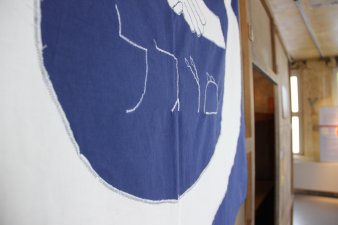 Eine blaue Fahne hängt an einer Holzwand. Aufgenäht ist ein weißer Kreis, mittig 2 Hände und ein hebräisches Wort.