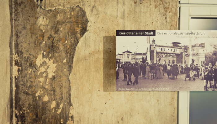 In der rechten Bildhäfte hängende Tafel mit einem Schwarz-Weiß-Foto, dass Stadtansicht mit Heil-Hitler-Tor und Menschenansammlung zeigt. Oben im Foto steht: Gesichter einer Stadt. Das nationalsozialistische Erfurt.In der linken Bildhälfte gelbe Wand mit alten Spuren.