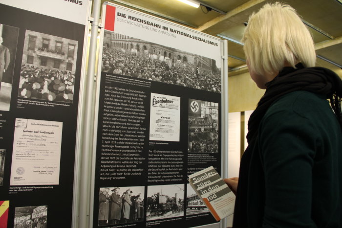 Junge weibliche Person, seitlich und leicht von unten fotografiert, steht vor einer schwarzen Tafel mit weißer Schrift und mehreren Abbildungen. Oben an der Tafel steht: Die Reichsbahn im Nationalsozialismus, Gleichschaltung und Anpassung. Junge Frau hält ein Faltblatt in der Hand mit lesbarem Text: Sonderzüge in den Tod.