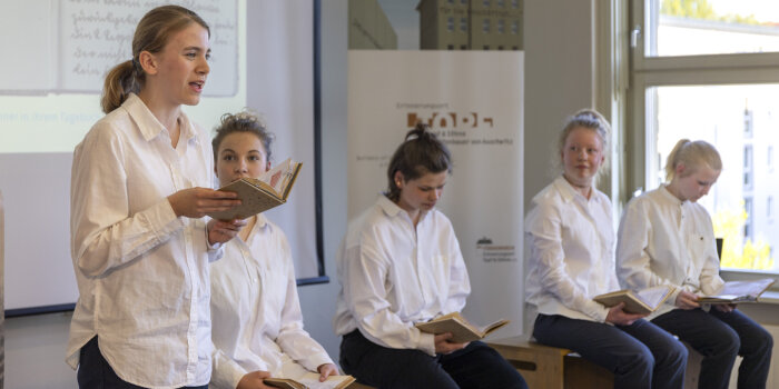 fünf Mädchen in weißen Blusen, eines von ihnen liest aus einem Buch vor