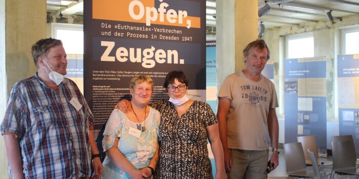 Vier Personen stehen vor einem Banner der Sonderausstellung "Täter, Oper, Zeugen"