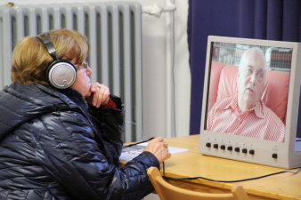 Farbfoto, eine Frau mit Kopfhörern sitzt vor einem Monitor, der einen älteren Mann zeigt