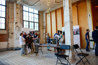 Farbfoto, Menschengruppe stehen um einen Tisch, auf dem drei Monitore mit Kopfhörern stehen