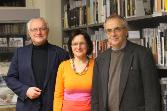 2 Männer und eine Frau stehen vor einem Bücheregal in einer Bibliothek.