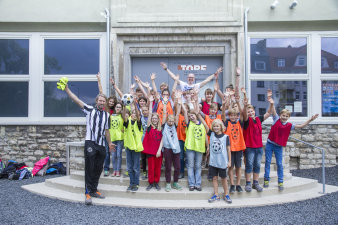 Farbfoto, Kinder stehen auf einer Eingangstreppe und werfen verschiedenfarbige Trikots in die Luft
