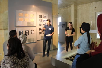 Ein junger Mann erklärt Besucherinnen und Besuchern etwas in einer Ausstellung