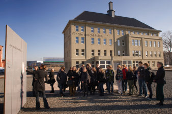 Im Vordergrund eine Gruppe von Jugendlichen, die zu einer weibliche Person links im Bild vor einer grauen Stele schaut. Im Hintergrund Gebäude mit Spitzdach