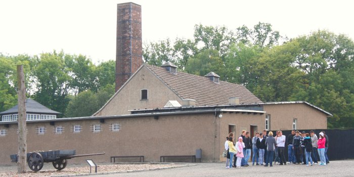 Graues verputztes Gebäude mit Spitzdach und großem Kaminschlot aus Backstein,vor dem Gebäude rechts eine größere Gruppe, links ein Handwagen