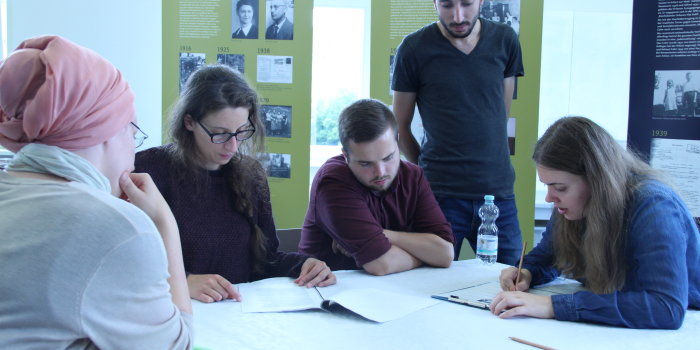 Fünf Personen arbeiten gemeinsam an einem Tisch, im Hintergrund ist eine Ausstellung zu sehen. 