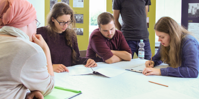 Mehre Personen arbeiten an einem Tisch und hinter ihnen stehen Ausstellungsbanner. 