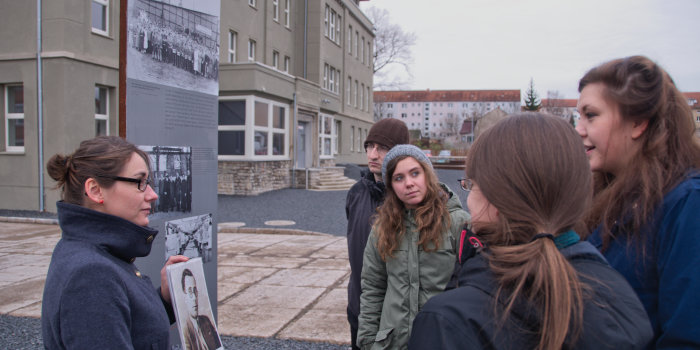 Eine Frau steht neben einer Informationsstele mit einem Blatt in der Hand und spricht mit vier Jugendlichen. 