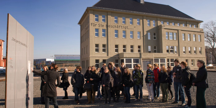 Im Vordergrund eine Gruppe von Jugendlichen, die zu einer weibliche Person links im Bild vor einer grauen Stele schaut.Im Hintergrund Gebäude mit Spitzdach