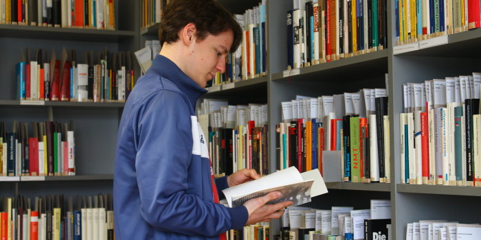 Ein junger Mann steht vor Bücherregalen, in seinen Händen ein Buch. Er liest. 