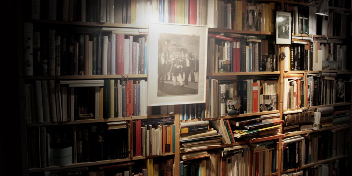 Gefüllter Bücherschrank nimmt gesamte Bildfläche ein,In der Bildmitte hängt am Bücherschrank ein historisches Foto mit Rahmen, Eine Lampe ist auf das Bild gerichtet und erleuchtet es.