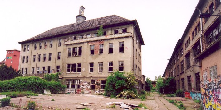 Ruine eines Hauses mit Spitzdach, leicht von der Seite fotografiert, Haus mit eingeschlagenen Fenstern, Graffiti und Pflanzenbewuchs, in der rechten Bildecke weitere Ruine eines größeren Gebäudes,Im Vordergrund größerer Vorplatz mit verschiedenem Gerümpel