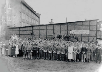 Menschen stehen in Reihe auf einem größeren Platz. Im Hintergrund sind Gebäude zu sehen.Schwarz-Weiß-Fotografie