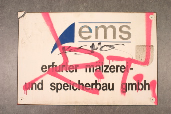 Weißes Firmenschild mit der Aufschrift: ems, erfurter mälzerei- und speicherbau gmbh Das Schild ist mit rotem und schwarzem Graffiti beschmiert.