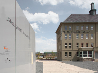 In der rechten Bildhälfte im Hintergrund: Hausansicht mit Schriftzug "Stets gern für Sie beschäftigt,..." an der HauswandIn der linken Bildhälfte im Vordergrund fünf sehr nahe nebeneinander stehende Einzelstelen mit Fluchtlinien zur Bildmitte. Selber Schriftzug über Einzelstelengrenze hinweg wie an der Hauswand 