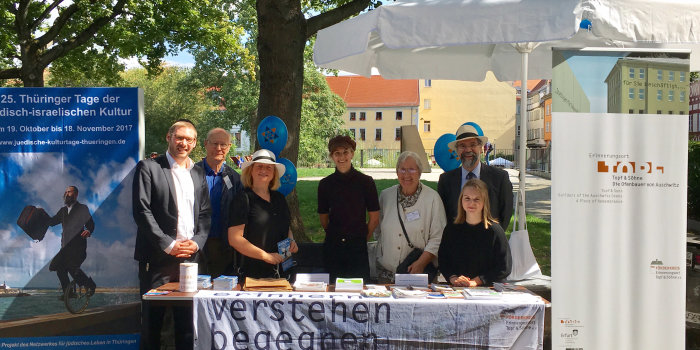 Farbfoto, sieben Personen stehen versammelt hinter einem Tisch, auf dem eine beschriftete Fahne liegt sowie unterschiedliche Druckerzeugnisse, ganz links und ganz rechts stehen zwei unterschiedliche Banner und ein Sonnenschirm, dazwischen blaue Luftballons