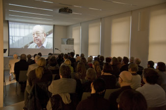 Publikum sitzt vor einer Leinwand, auf der ein älterer Mann zu sehen ist