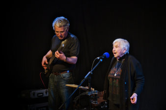Alte singende Frau am Mikrofon, neben ihr älterer Mann an der E-GitarreSchwarzer Hintergrund