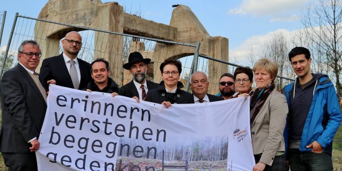 Farbfoto, Personengruppe hält Banner mit Schriftzug erinnern, verstehen, begegnen, gedenken, im Bildhintergrund Reste einer Ruine