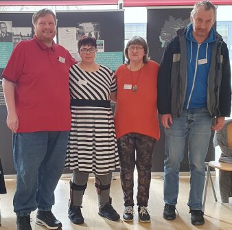 Zwei Männer und zwei Frauen stehen in einer Reihe vor Geschichtsaufstellern und schauen gemeinsam in die Kamera.