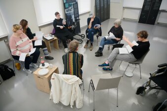 Sieben Leute sitzen in einem Stuhlkreis zusammen, halten Skripte in den Händen und besprechen etwas.