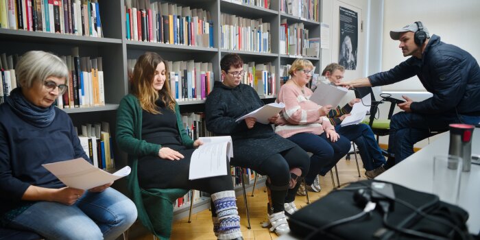 Fünf Leute sitzen in einer Stuhlreihe und halten ein Skript, vor ihnen sitzt ein Aufnahmeleiter mit Mikrofon.