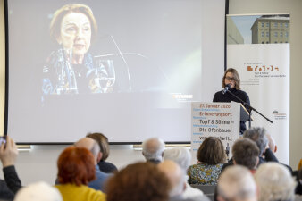Frau an Mikrofon, die eine Rede vor Publikum hält. Im Hintergrund eine Frau auf der Leinwand